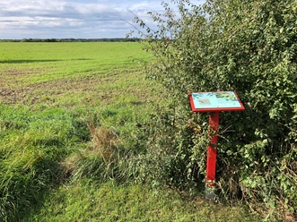 Een rood informatiebord staat aan de rand van het veld, omringd door struiken en groene weiden.