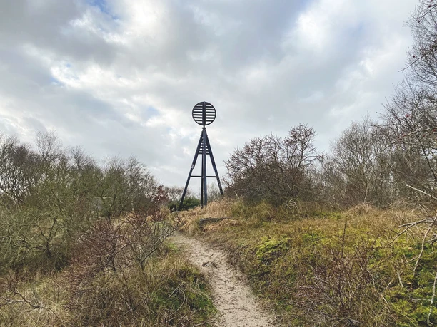 Die Kugelbake thront als hölzernes Seezeichen am Horizont, umgeben von Dünen und Küstengras.The Kugelbake stands as a wooden sea marker on the horizon, surrounded by dunes and coastal grass.