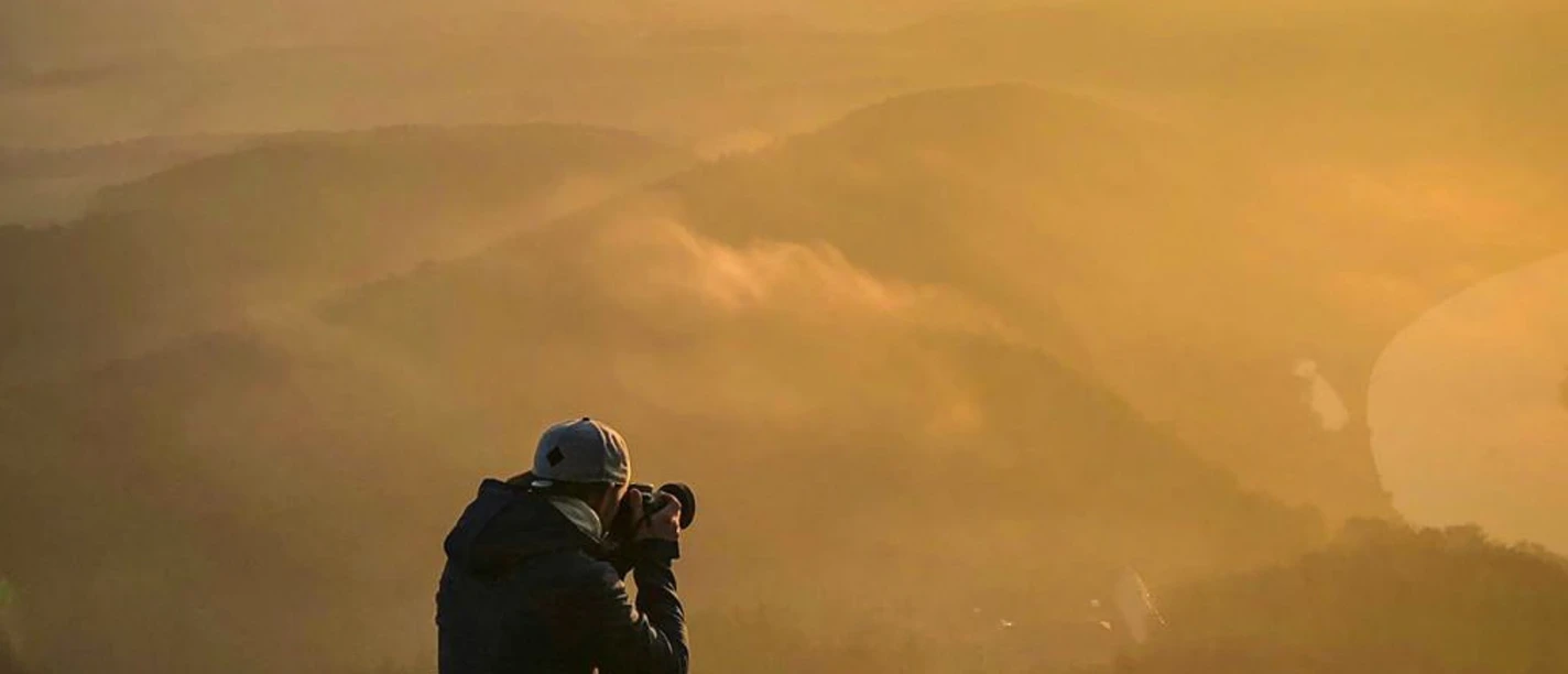titelbild_33__Fotografieren_in_der_Sächsischen_Schweiz_Lilienstein_©_Sebastian_Rodenstock.jpeg Mensch fotografiert bei Sonnenaufgang von einer Klippe aus die nebelverhangene Landschaft.