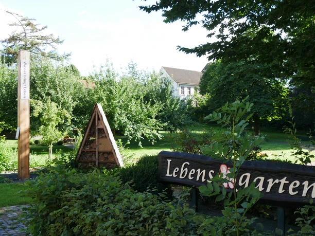Holzpfahl mit Bibelzitat im grünen Lebensgarten vor einem Haus, umgeben von Bäumen und Pflanzen.