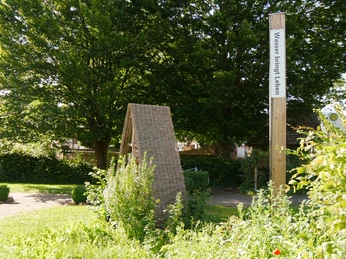 Bibel-Pfahl Amelunxen Holzpfosten mit Bibelvers "Wasser bringt Leben" neben einer geflochtenen Strukturskulptur im Grünen.