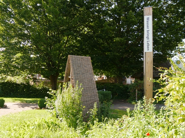 Bibel-Pfahl Amelunxen Holzpfosten mit Bibelvers "Wasser bringt Leben" neben einer geflochtenen Strukturskulptur im Grünen.