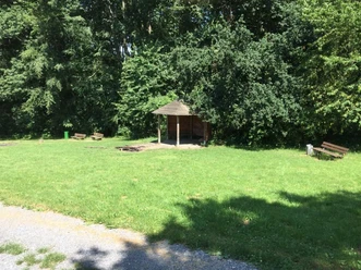 Ein hölzerner Unterstand auf einer Grasfläche, umgeben von Bäumen, mit mehreren Holzbänken.