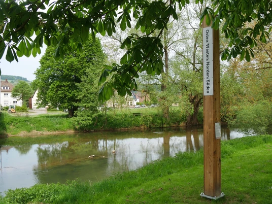 Bibel-Pfahl-Bruchhausen Holzpfeiler mit Bibelzitat an einem ruhigen Flussufer, umgeben von Bäumen und grüner Landschaft.