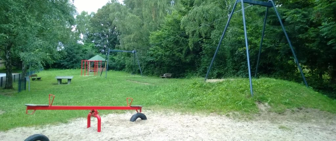 Spielplatz mit roter Wippe und Rutsche, umgeben von üppigem Grün und schattenspendenden Bäumen.