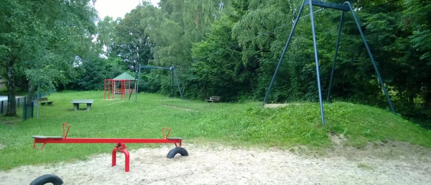 Kinderspielplatz an der Osterholzer Straße Spielplatz mit roter Wippe und Rutsche, umgeben von üppigem Grün und schattenspendenden Bäumen.
