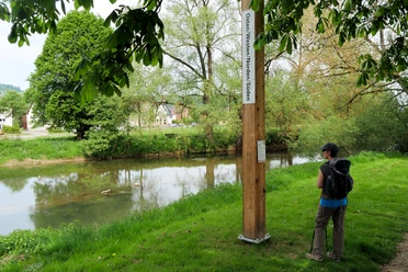 Ein Holzpfahl mit Himmelsrichtungen am Ufer eines ruhigen Flusses, ein Wanderer mit Rucksack schaut.