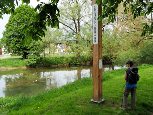 Bibel-Pfahl-Bruchhausen Ein Holzpfahl mit Himmelsrichtungen am Ufer eines ruhigen Flusses, ein Wanderer mit Rucksack schaut.