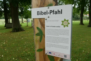 Bibel-Pfahl Brakel Holzpfahl im Grünen mit Beschilderung zu einem Bibelzitat, umgeben von hohen Bäumen.