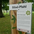 Bibel-Pfahl Brakel Holzpfahl im Grünen mit Beschilderung zu einem Bibelzitat, umgeben von hohen Bäumen.