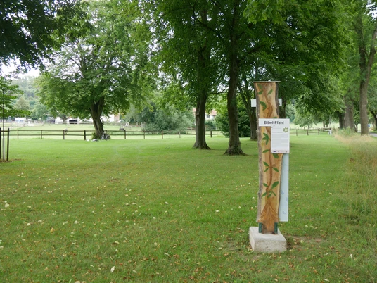 Bibel-Pfahl Brakel Ein Holzpfahl mit Bibelversen steht auf einer grünen Wiese, umgeben von hohen Bäumen.