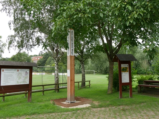 Bibel-Pfahl Riesel mit Infotafeln, von Bäumen umgeben, nahe einem Sportfeld in ländlicher Umgebung.