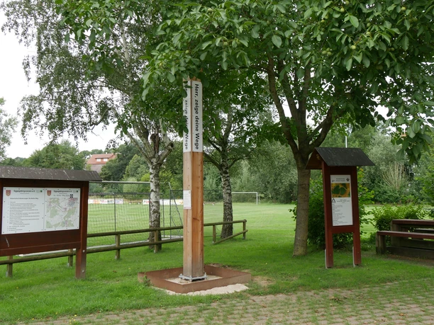 Bibel-Pfahl Riesel mit Infotafeln, von Bäumen umgeben, nahe einem Sportfeld in ländlicher Umgebung.