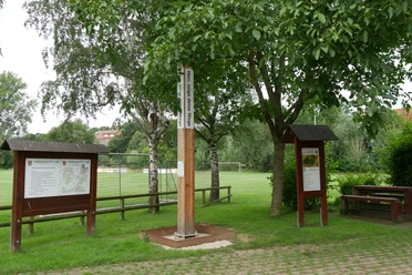 Bibel-Pfahl Riesel Bibel-Pfahl Riesel mit Infotafeln, von Bäumen umgeben, nahe einem Sportfeld in ländlicher Umgebung.