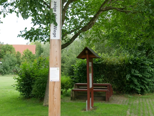 Ein hölzerner Bibelpfahl steht auf einem gepflasterten Weg, umgeben von Bäumen und grünen Büschen.