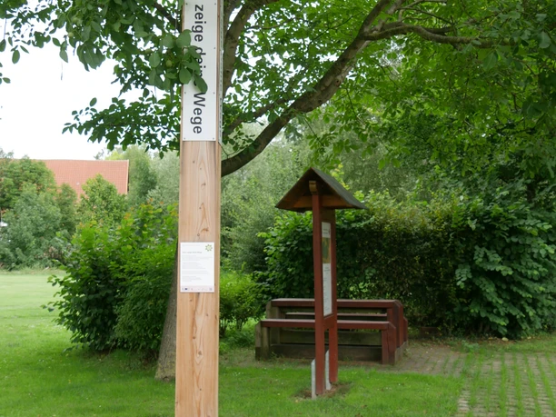 Bibel-Pfahl Riesel Ein hölzerner Bibelpfahl steht auf einem gepflasterten Weg, umgeben von Bäumen und grünen Büschen.