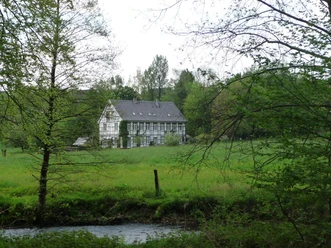 Brucher Kotten von der Itterseite aus Historisches Fachwerkgebäude am Fluss, umgeben von grüner Wiese und Bäumen im Hintergrund.