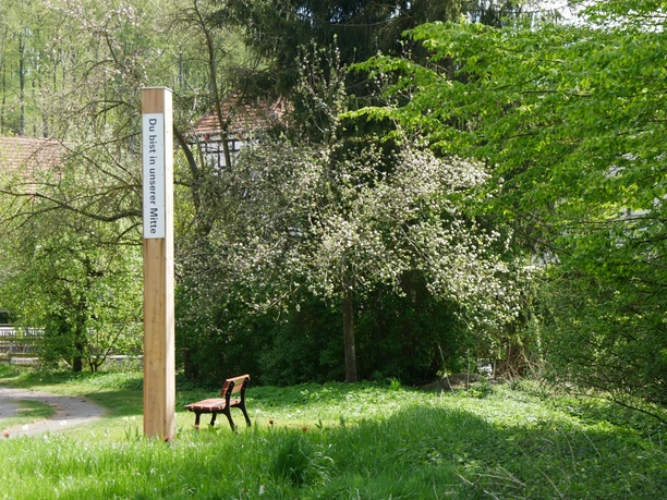 Ein hölzerner Pfahl mit der Inschrift "Du bist in unserer Mitte" in einer grünen, bewaldeten Umgebung.