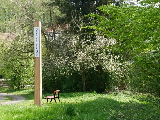 Bibel-Pfahl-Rheder Ein hölzerner Pfahl mit der Inschrift "Du bist in unserer Mitte" in einer grünen, bewaldeten Umgebung.