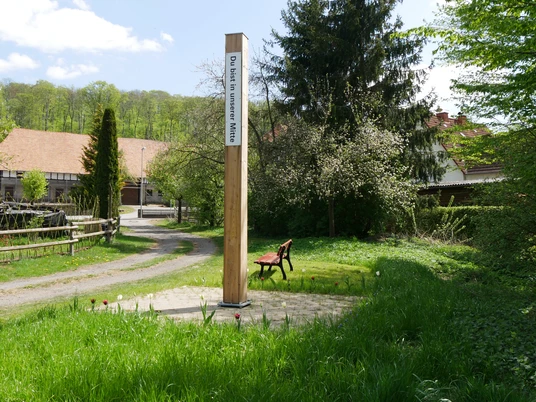 Bibel-Pfahl-Rheder (5).jpg Ein hölzerner Pfahl mit Schriftzug auf einer Wiese, umgeben von Bäumen und einem Weg.