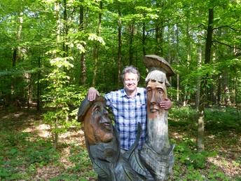 im Waldklassenzimmer Ein Mann steht lächelnd neben einer hölzernen Skulptur von zwei Figuren im grünen Waldklassenzimmer.