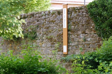 Bibel-Pfahl Gehrden Steinmauer mit angelehntem Holzpfahl und Schild; umgeben von grüner Vegetation.