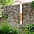 Bibel-Pfahl Gehrden Steinmauer mit angelehntem Holzpfahl und Schild; umgeben von grüner Vegetation.