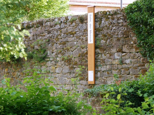 Bibel-Pfahl Gehrden Steinmauer mit angelehntem Holzpfahl und Schild; umgeben von grüner Vegetation.