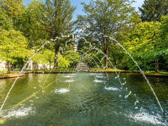Park von Schloss Landsberg nahe Essen Wasserspiele in einem Park, umgeben von grünen Bäumen und einem steinernen Bauwerk im Hintergrund.