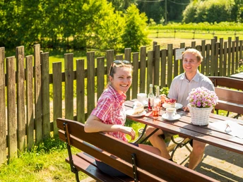 Stärkung in den Hofcafés nahe des neanderland STEIGs Ein Paar genießt Kaffee und Kuchen in einem sonnigen Garten, umgeben von grünem, ländlichem Ambiente.