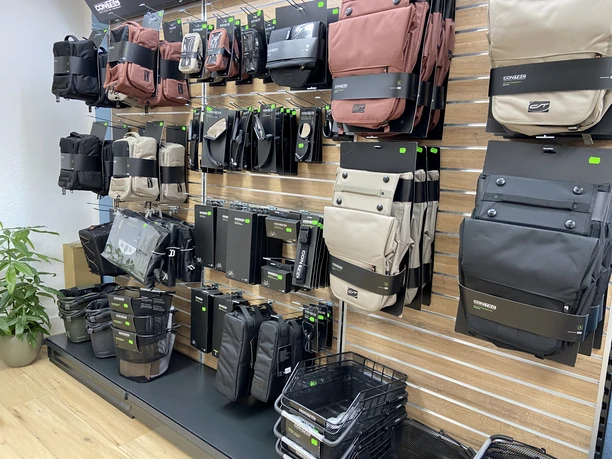Bikefactory5.jpeg Verschiedene Fahrradtaschen sind in einem Geschäft an einer Wand präsentiert. Links steht eine Pflanze. Der Boden ist aus Holz.