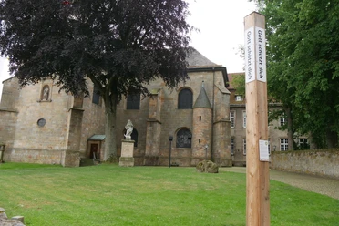 Bibel-Pfahl Willebadessen Kirche mit Fachwerkmauer in Willebadessen. Vordergrund: Holzpfahl mit Bibelzitat, grüner Rasen.