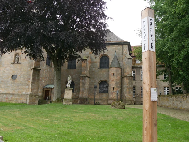 Bibel-Pfahl Willebadessen Kirche mit Fachwerkmauer in Willebadessen. Vordergrund: Holzpfahl mit Bibelzitat, grüner Rasen.