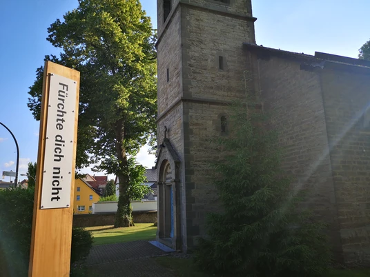 Bibel-Pfahl Peckelsheim Kirche mit markantem Turm, umgeben von Bäumen, daneben ein Pfahl mit der Aufschrift "Fürchte dich nicht".