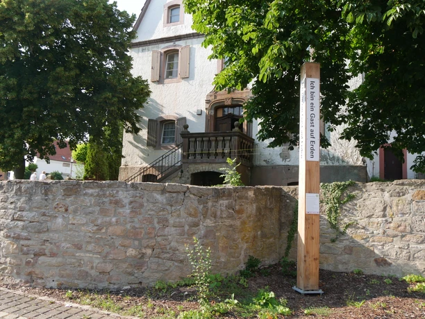 Ein Bibel-Pfahl in Bonenburg steht vor einem Haus, von Bäumen umgeben und mit einer Steinmauer.