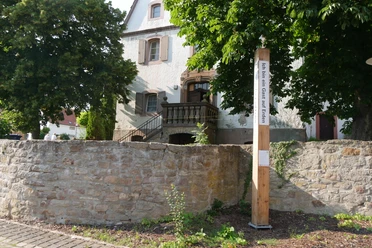 Bibel-Pfahl Bonenburg Ein Bibel-Pfahl in Bonenburg steht vor einem Haus, von Bäumen umgeben und mit einer Steinmauer.