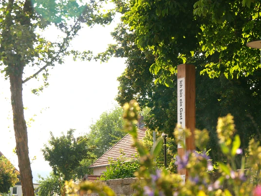 Ein Holzpfahl mit Bibelzitat, umgeben von blühenden Pflanzen, unter schattenspendenden Bäumen.