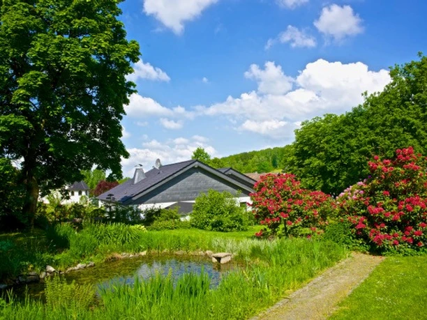 Wilkenroth Ein idyllischer Garten mit Teich, bunten Blumen und grünen Bäumen vor einem Landhaus unter blauem Himmel.