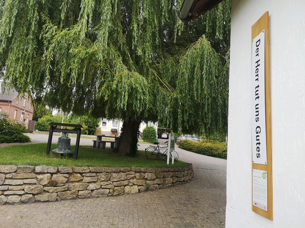 Der Bibel-Pfahl Rimbeck zeigt einen Schriftzug neben einer Glocke unter einem großen, hängenden Baum.