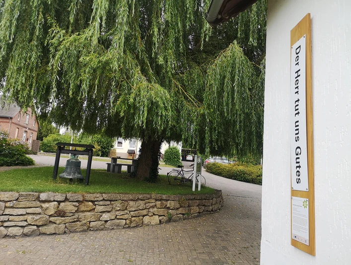 Der Bibel-Pfahl Rimbeck zeigt einen Schriftzug neben einer Glocke unter einem großen, hängenden Baum.