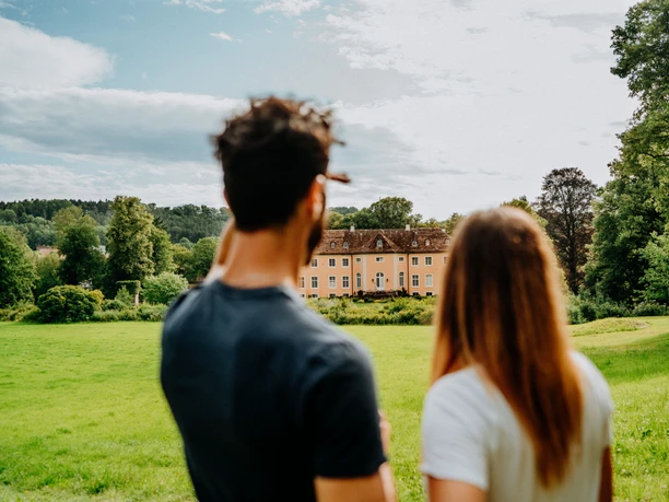 Blick auf Schloss Rheder in Brakel Zwei Personen betrachten die Fassade des gelben Schlosses Rheder in Brakel, umgeben von Grünflächen.
