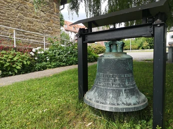 Glocke unter einem Schutzdach im Freien, umgeben von Gras und Blüten vor einem steinernen Bauwerk.
