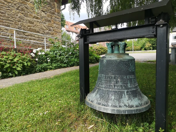 Glocke unter einem Schutzdach im Freien, umgeben von Gras und Blüten vor einem steinernen Bauwerk.