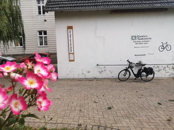 Bibel-Pfahl Rimbeck Elektro-Tankstelle vor einer Hauswand, rechts ein Fahrrad; im Vordergrund rosa Blumen.