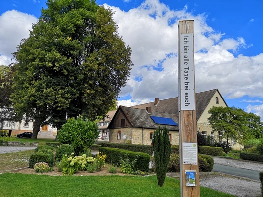 Pfahl mit Bibelzitat "Ich bin alle Tage bei euch" vor ländlicher Kirche und blauem Himmel.