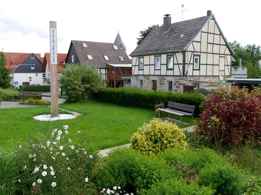 Bibel-Pfahl Germete Dorfplatz mit gepflegtem Garten, traditionellem Fachwerkhaus und Bibel-Pfahl im Vordergrund.