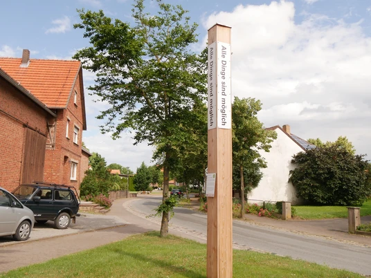 Holzpfahl mit Bibelzitat in Körbecke, umgeben von Wohnhäusern, Bäumen und einer Straße.