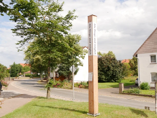 Ein hoher Holzpfahl mit Bibelversen steht in einem grünen Vorort, umgeben von Häusern.