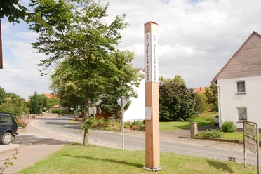 Bibel-Pfahl 19 - Körbecke Ein hoher Holzpfahl mit Bibelversen steht in einem grünen Vorort, umgeben von Häusern.