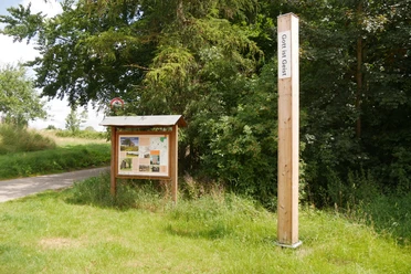 Bibel-Pfahl 20 - Borgentreich Ein hoher Holzpfahl mit der Aufschrift "Gott ist Geist" steht neben einer Infotafel am Wegesrand.
